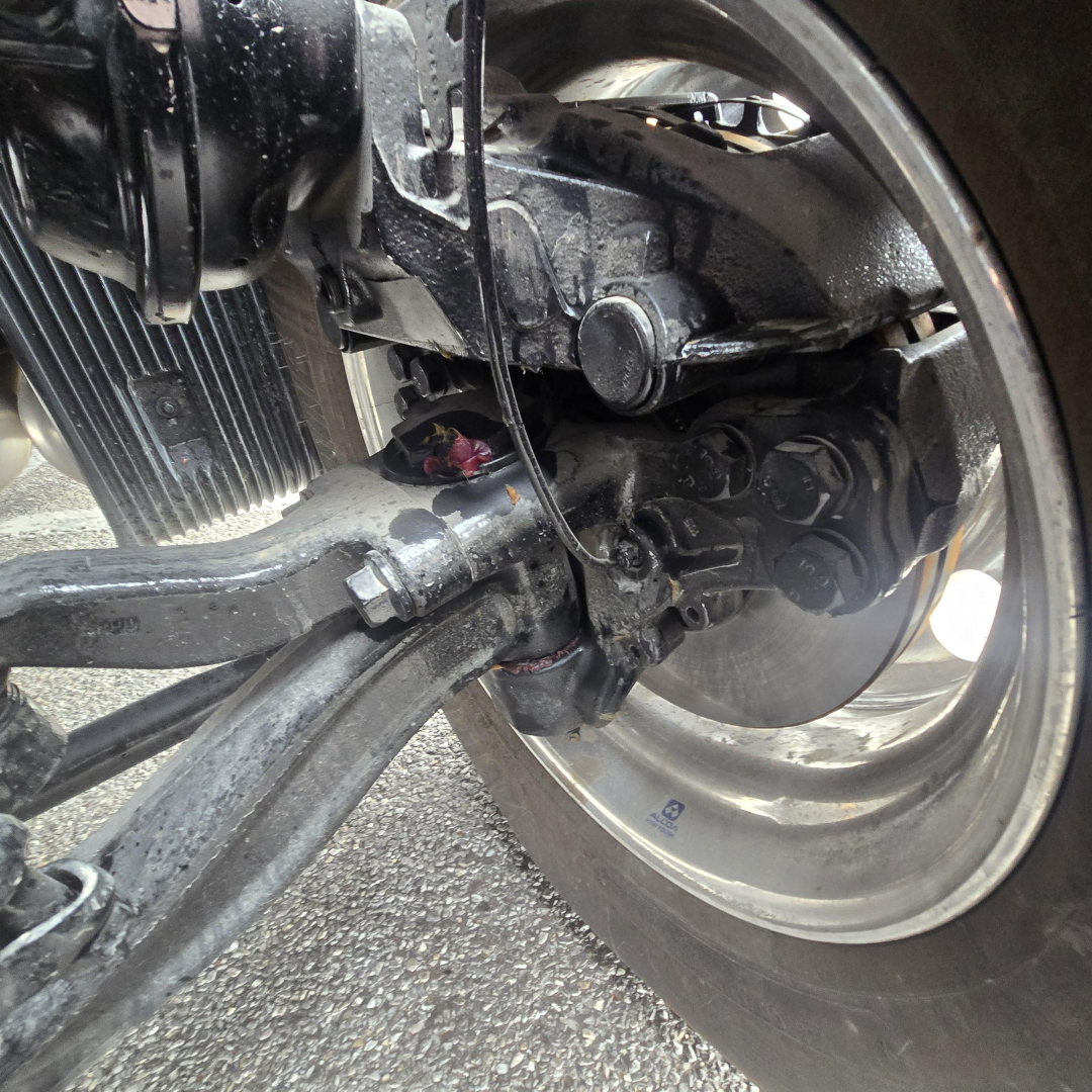 Axle and suspension components of a Mack PR64T76M truck showing wheel hub, brake drum, and linkage with metallic finish.