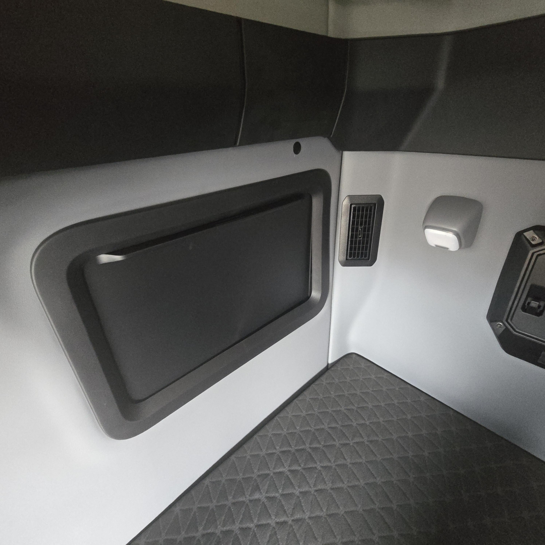 Trucker cab interior paneling with a storage hatch and ventilation vent on the side of a Mack PR64T76M truck interior.