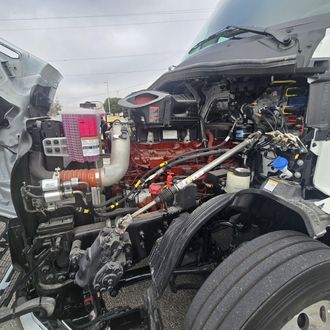 Engine bay of a new 2026 Mack PR64T76M truck showing exposed diesel engine, intake, hoses, and front suspension; visible components include air intake, belts, wiring, and coolant reservoir.