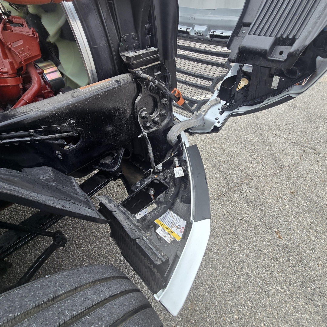 Truck engine bay area with hood open showing chassis and front bumper, side view of cab access and warning sticker.