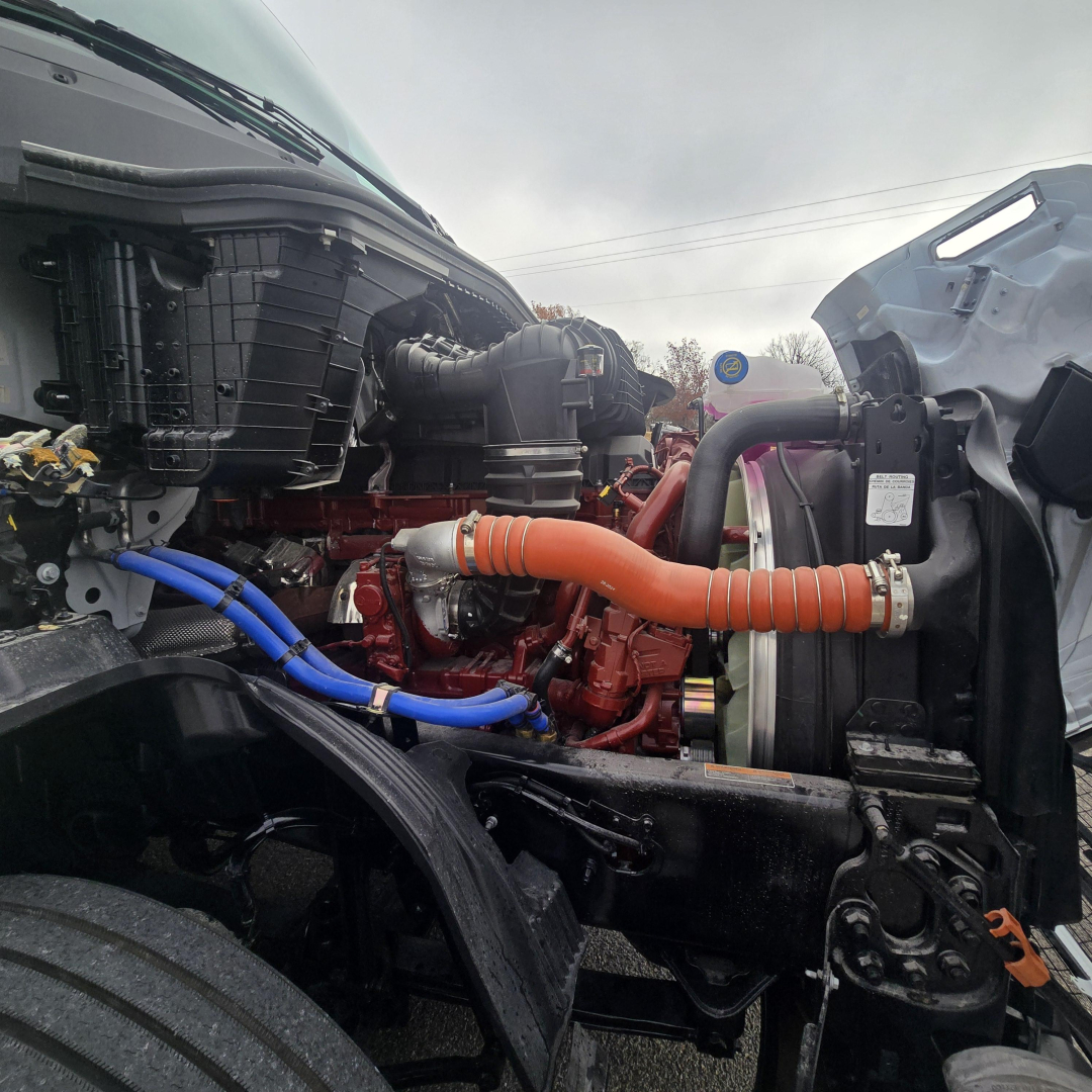 Truck engine bay view showing red engine, blue and orange hoses, and front radiator area; Mack PR64T76M engine components visible.