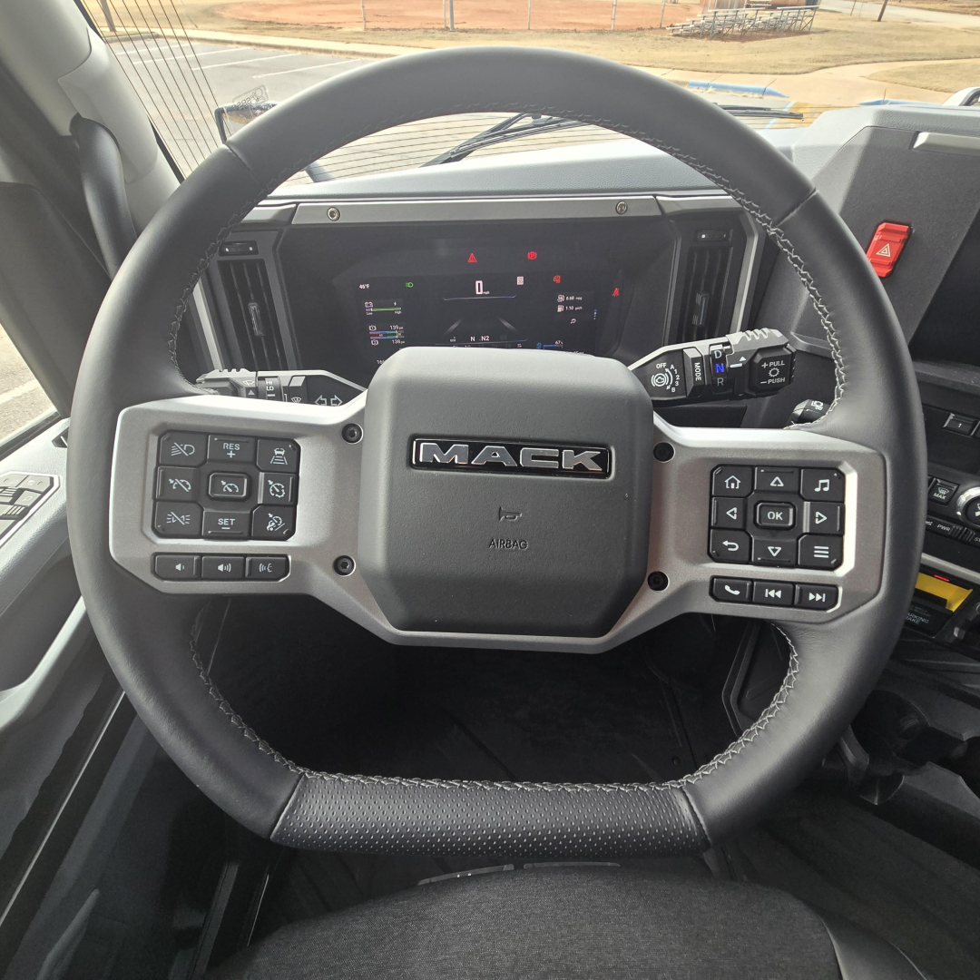 Mack truck steering wheel with dashboard controls in view. Center badge reads MACK. Left and right control clusters on steering wheel show multiple buttons. Instrument panel behind displays digital gauges.