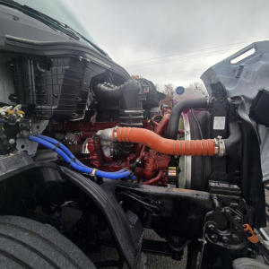 Truck engine bay view showing red engine, blue and orange hoses, and front radiator area; Mack PR64T76M engine components visible.
