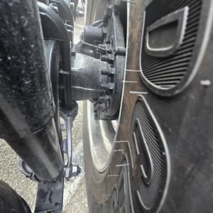 Truck wheel and suspension components with heavy-duty hub, brake drum, and tire profile visible; close-up view of the front axle area.