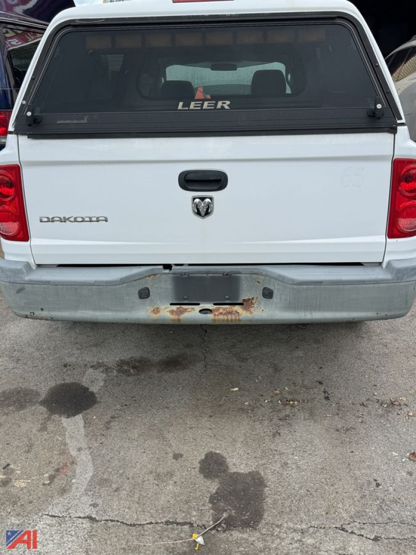 Isolated rear view of a white 2006 Dodge Dakota pickup with cap, showing tailgate with Dodge emblem, RAM logo, and rusted bumper area.