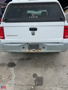 Isolated rear view of a white 2006 Dodge Dakota pickup with cap, showing tailgate with Dodge emblem, RAM logo, and rusted bumper area.