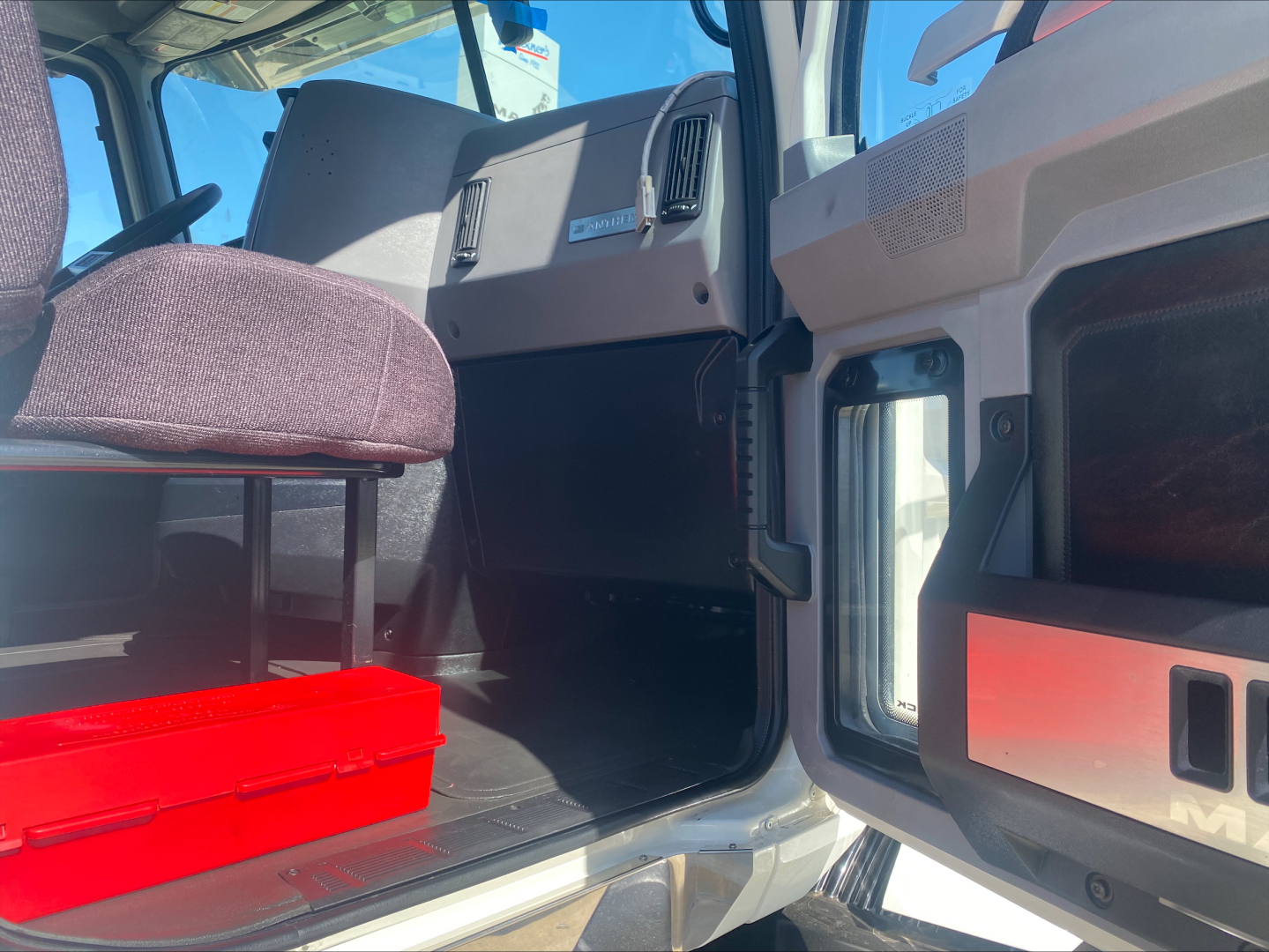 Truck cab interior showing entry step and dashboard area of a Mack AN64T with a red toolbox under the seat and VIN/stock info not visible inside the cab.
