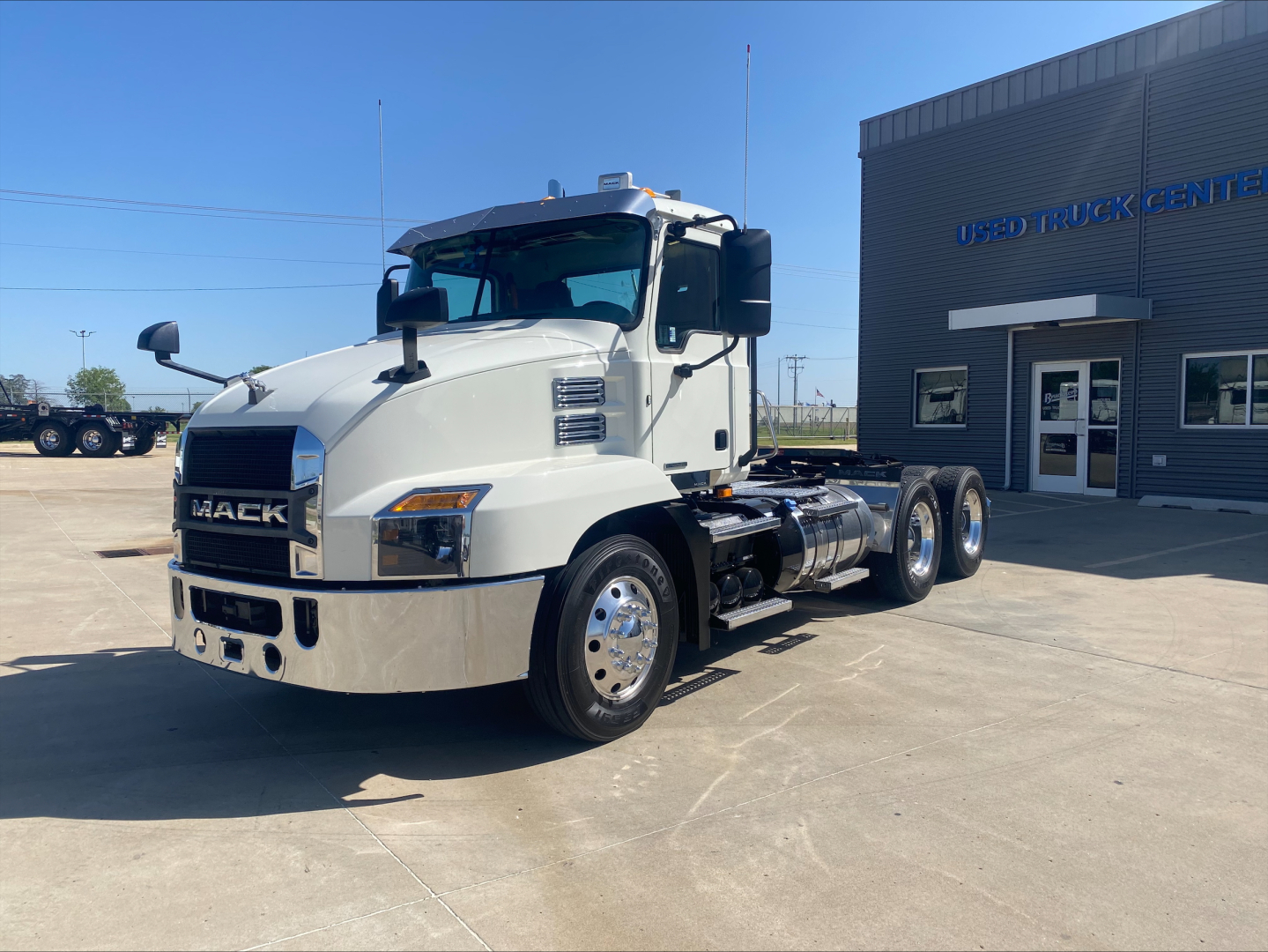 Mack AN64T conventional daycab truck in showroom lot showing front grille with Mack emblem and dual axles, white cab, chrome bumper, Used Truck Center building in background.