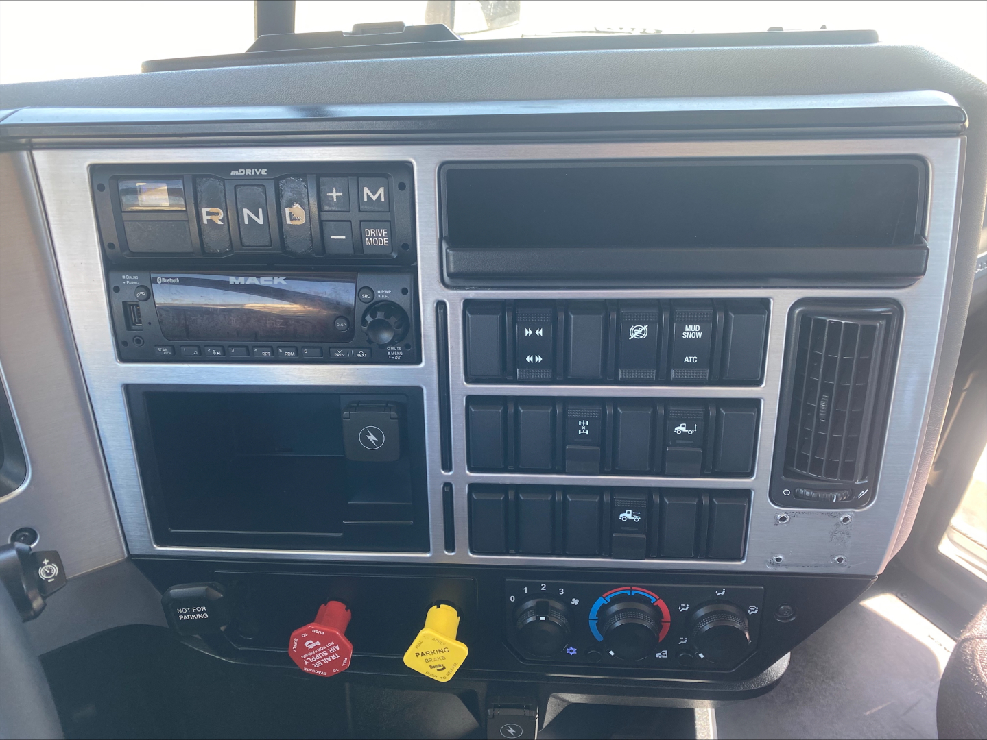 Dashboard view of a Mack AN64T showing gear selector, radio, climate controls, and various switches; prominent red hood release, yellow trailer brake knob, and round climate dial.