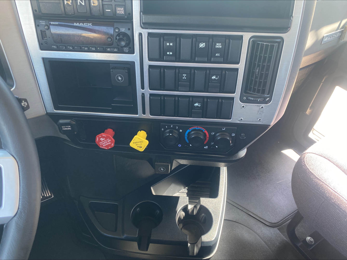 Dashboard of a Mack AN64T conventional daycab showing control panel with red and yellow knobs, climate dials, cup holders, and lower storage area.
