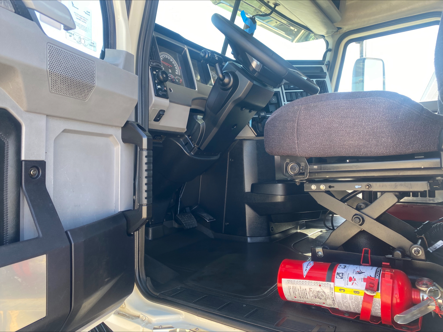 Mack AN64T truck interior cab view showing driver's seat, steering wheel, dashboard, and a red fire extinguisher mounted under the seat area.