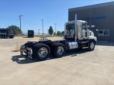 2021 Mack AN64T truck chassis side view showing empty fifth wheel and dual rear axles; cab with exhaust stack, fuel tanks, and wheels visible; dealership building in background.