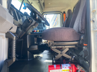 Interior of a 2021 Mack AN64T daycab front cabin showing driver seat and steering wheel with controls, instrument panel, and cabin floor; red fire extinguisher mounted near seat.