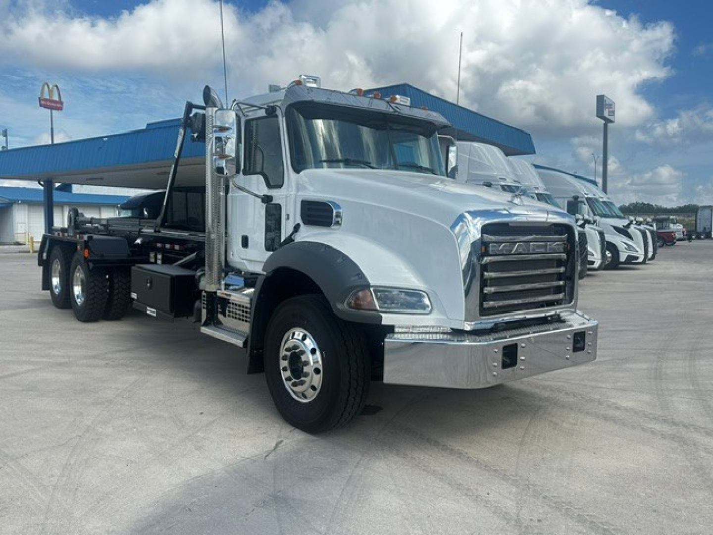 2026 Mack GR64B roll-off truck in light gray with chrome front bumper and black flatbed; multiple wheels visible; dealership lot in background.
