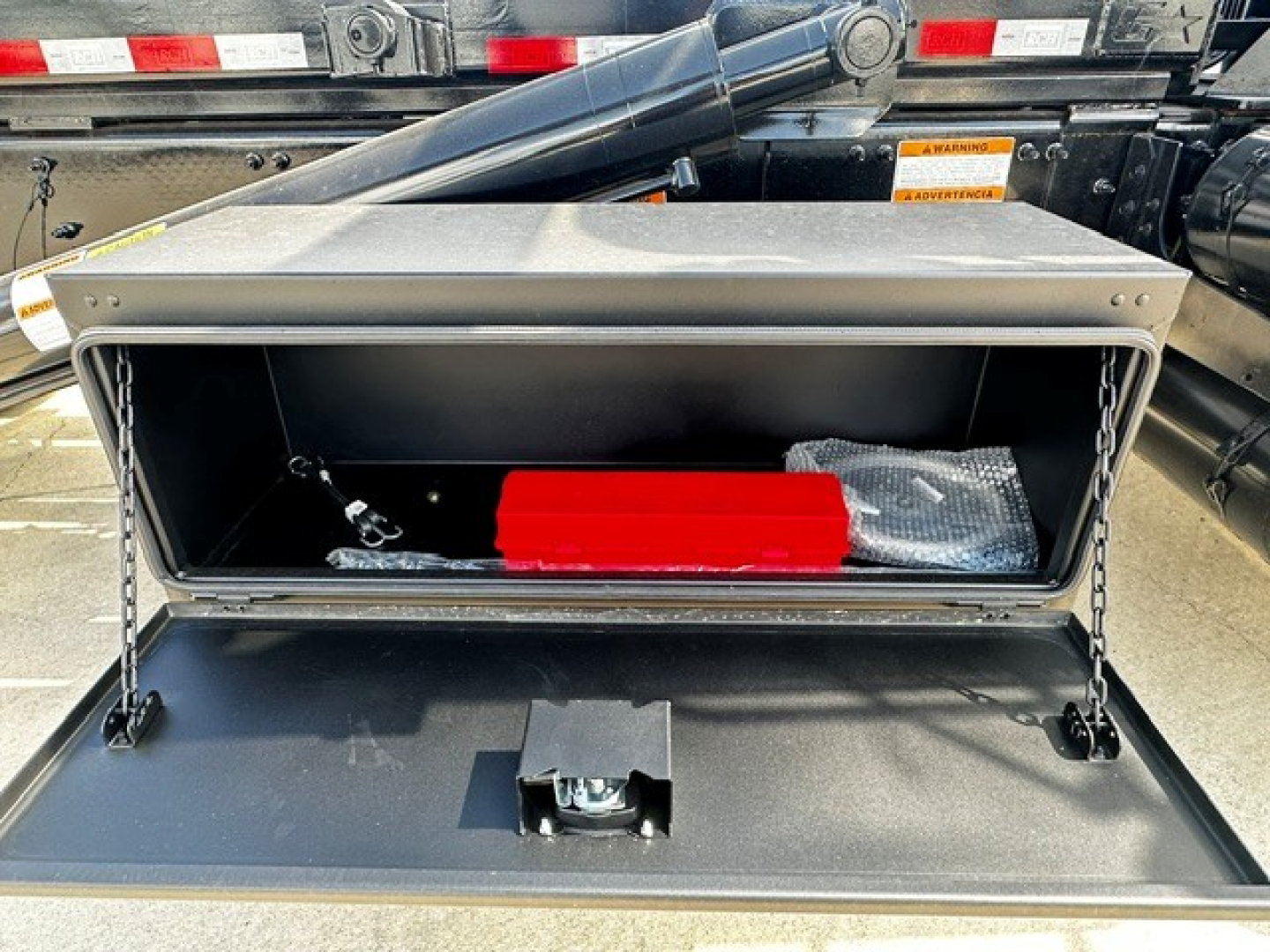 Truck component view showing an open storage compartment on a 2026 Mack GR64B roll-off truck with a red tool, metal frame, and hydraulic components in the background.