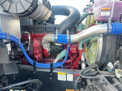 Mack GR64B engine bay with red engine block, blue hoses, silver intake pipe, purple coolant reservoir, and warning labels visible.