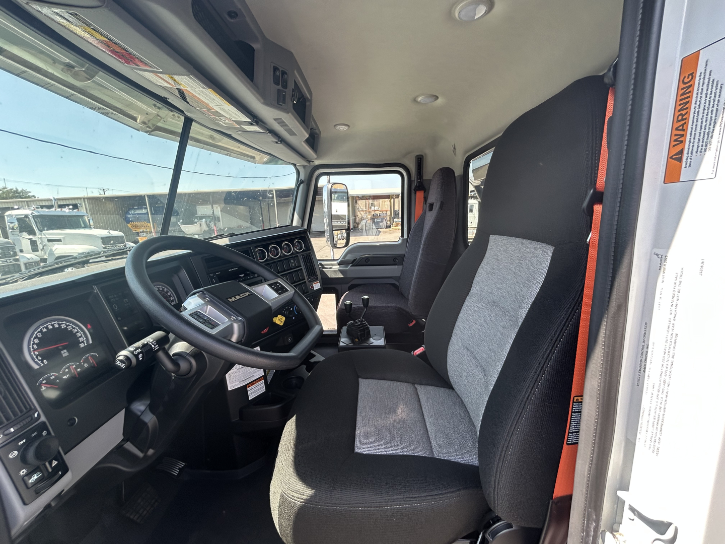 Truck cab interior of a 2026 Mack GR64B rolloff showing steering wheel, dashboard with gauges, driver and passenger seats, center console, and door frame with warning sticker.