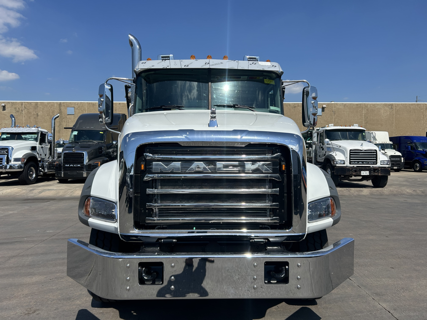 Front view of a white Mack truck with large chrome grille and bumper. Hood ornament, side mirrors, and other trucks visible in background.