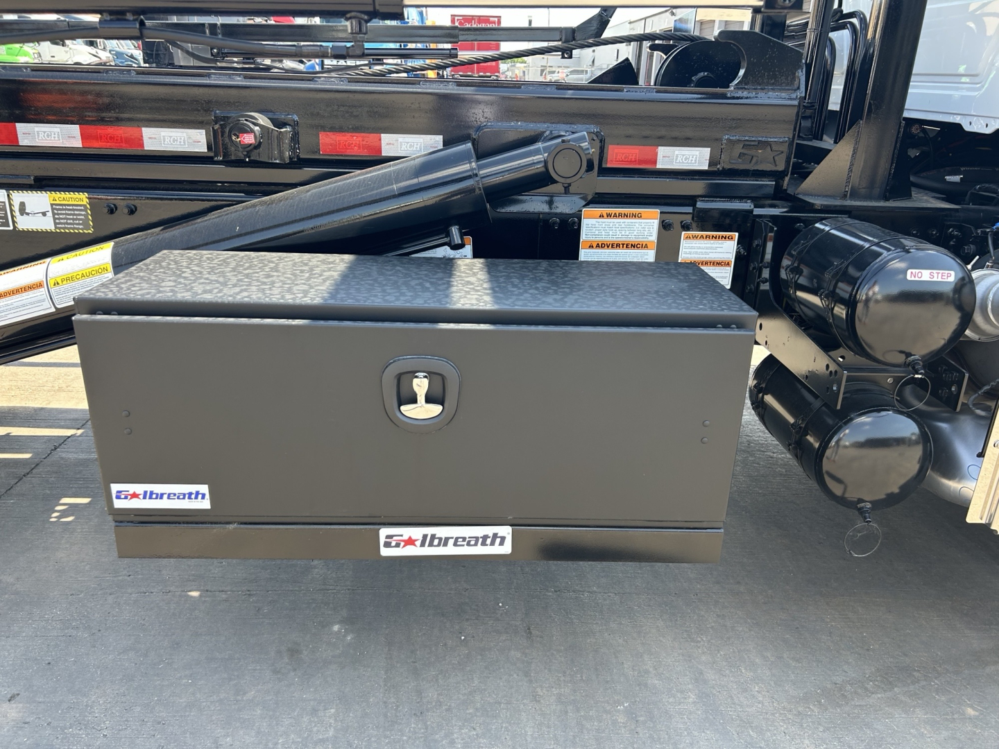 Rear view of a Mack GR64B roll-off truck showing a black metal toolbox with a silver handle, hydraulic arm, and Breth labels.