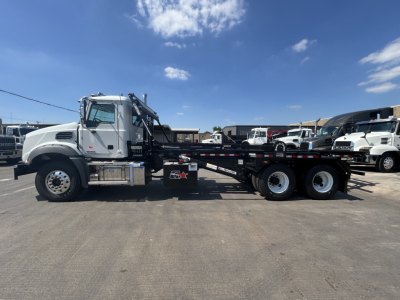 Truck side view of a 2026 Mack GR64B roll-off truck with a long flatbed and multiple rear axles, chrome cab, and hydraulic machinery mounted behind the cab.