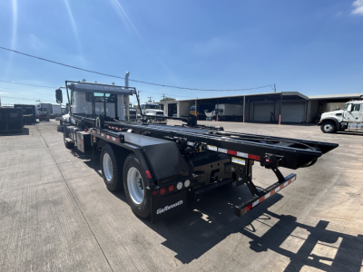2026 Mack GR64B roll-off truck in a yard, side and rear view showing flatbed and rear underride ramp, with dual rear axles and Mack branding.