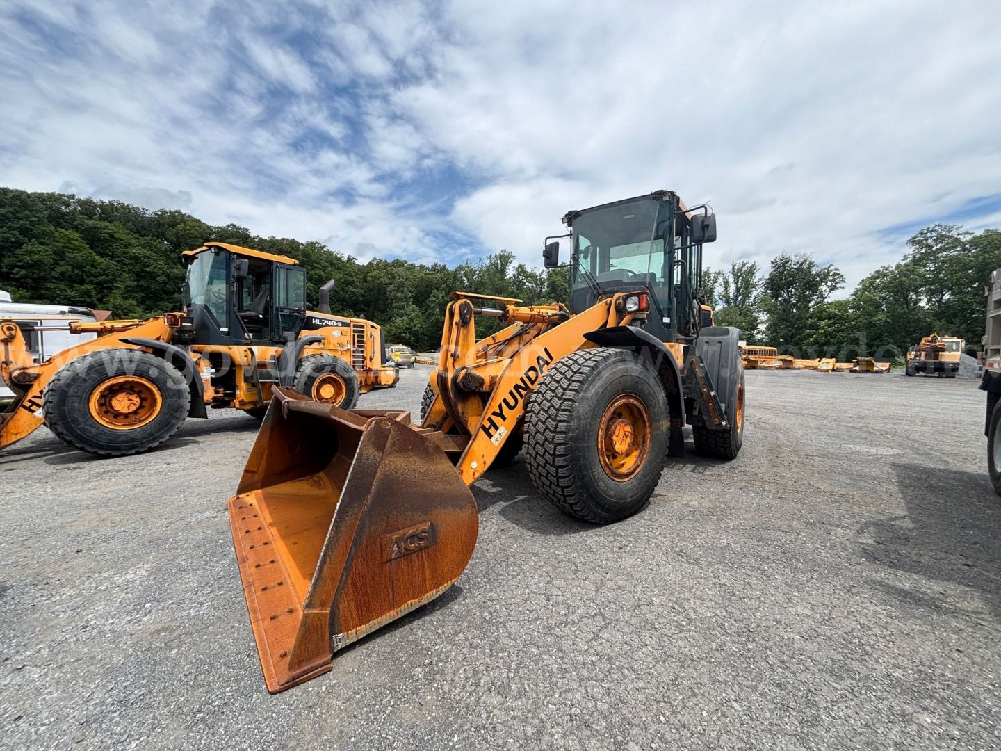 Hyundai HL740-9 wheel loader in a yard with a front bucket attachment, other loaders in background.