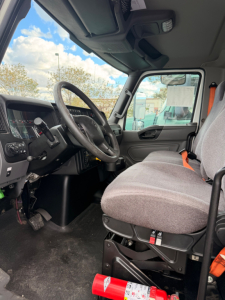 Freightliner M2-106 cab interior showing steering wheel, dashboard, gray fabric seats, and driver-side area with orange seat belt.