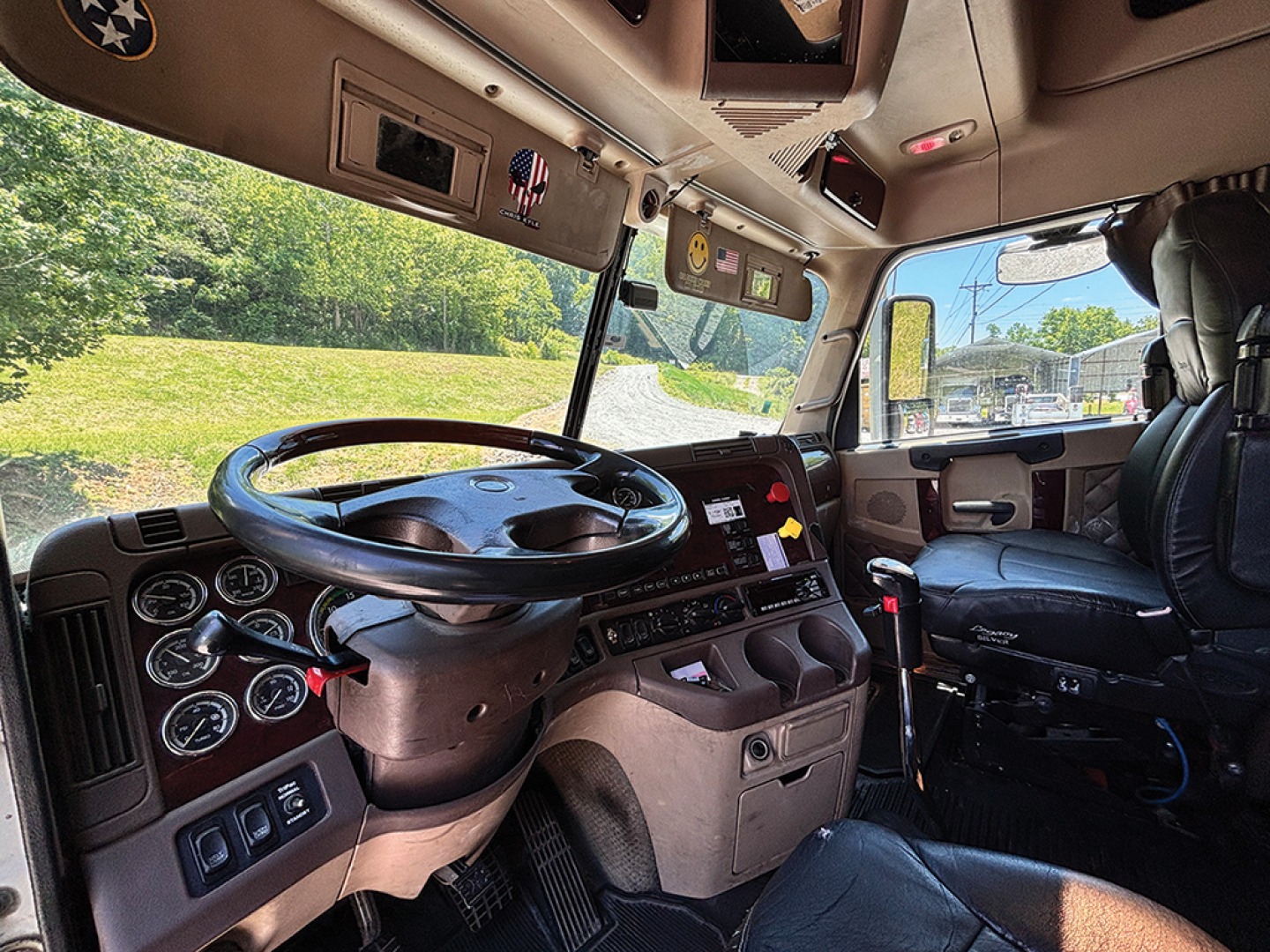 Freightliner Coronado truck cab interior showing steering wheel, dashboard gauges, and driver seat from the passenger side. Visible windows and sunlit cab.