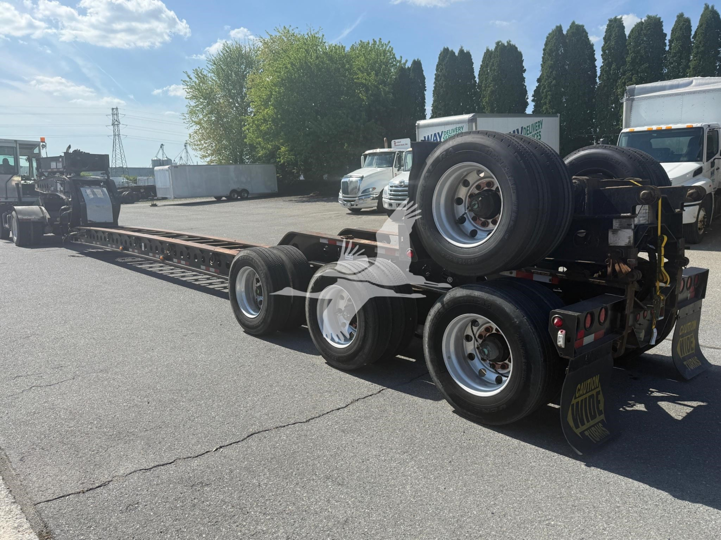 Trailer showing a long lowboy with multiple wheels and a detached spare tire on the rear; metal frame and axles visible; outdoor lot with trucks in background.
