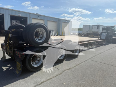 Trailer with two rear axles and spare tires, flatbed lowboy trailer in an outdoor lot. Visible text on yellow plate: 'X' and there are wheel rims and a storage building in background.
