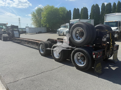 Trailer showing a long lowboy with multiple wheels and a detached spare tire on the rear; metal frame and axles visible; outdoor lot with trucks in background.
