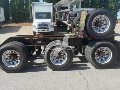 Lowboy trailer frame with tandem axles and a single wheel on the far side; spare wheel mounted on the bed and a white cab truck in the background. Visible text includes no legible labels on the trailer, but the setting shows a used trailer 