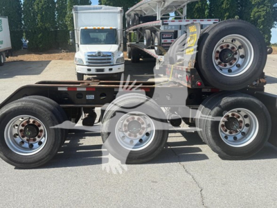 Trailer frame showing three axles with wheels, spare wheel, and a white cab in background; no visible labels legible.