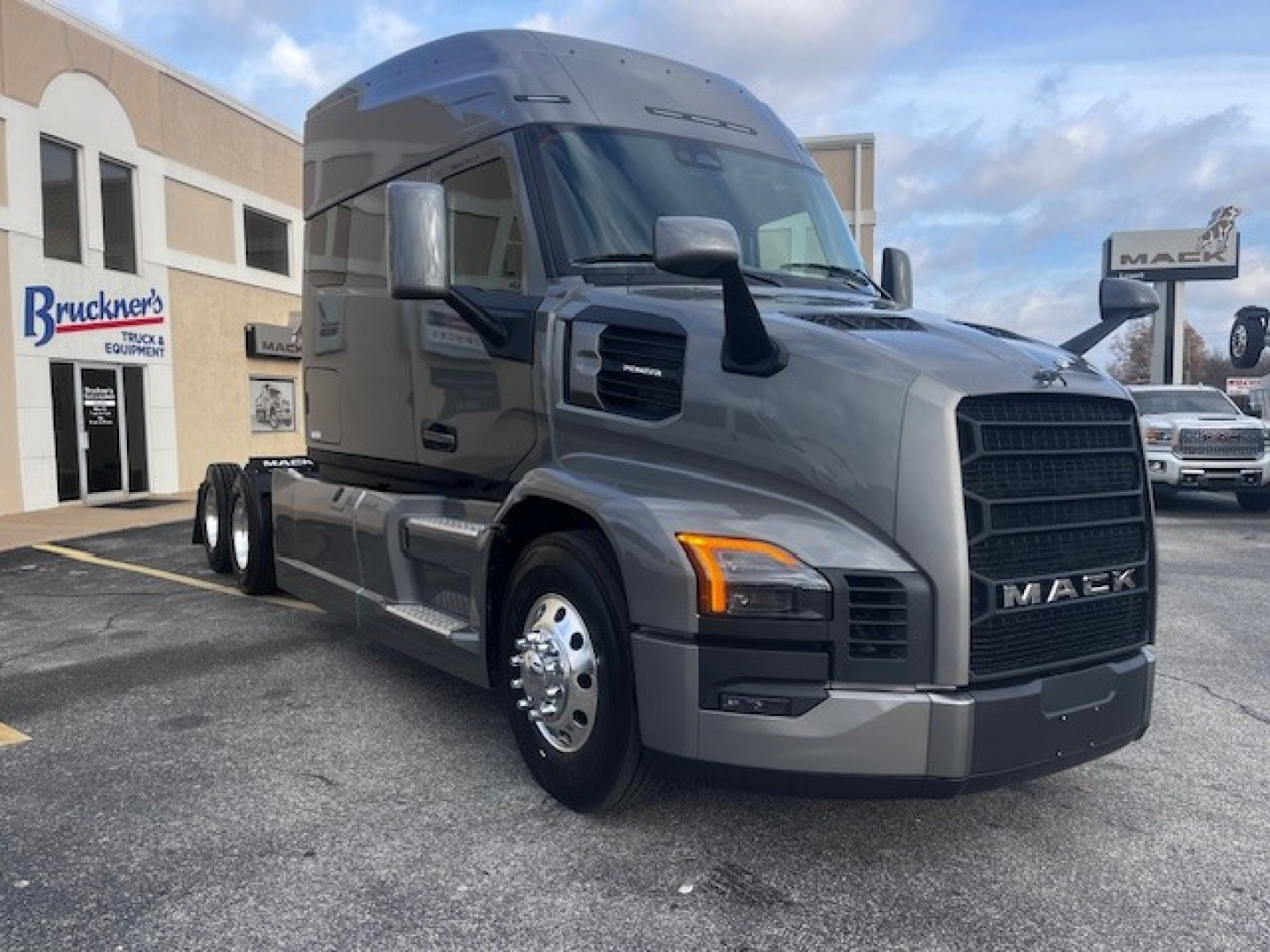 2026 Mack PR64T76M sleeper truck in showroom gray front-left view showing Mack grille and cab, with dealer lot in background.