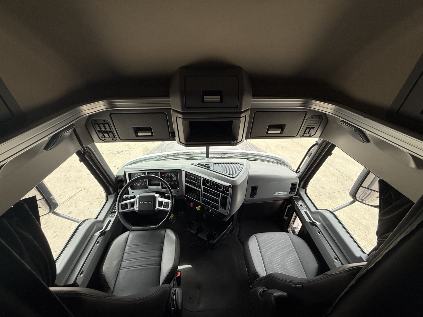 Truck cab interior of a 2023 Mack AN64T70 from a high interior perspective showing steering wheel, dashboard, captain chair, and passenger seat.