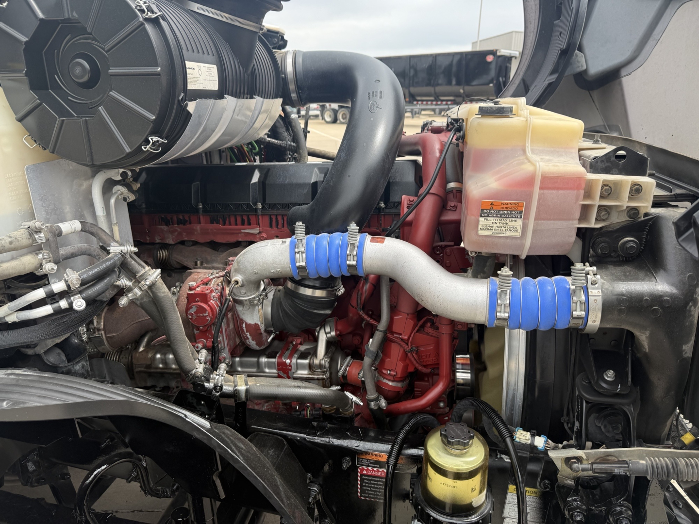Engine bay showing Cummins/Detroit style diesel with coolant reservoir, hoses, and metal brackets in a Mack AN64T70 truck engine compartment.