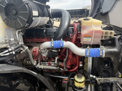 Engine bay showing Cummins/Detroit style diesel with coolant reservoir, hoses, and metal brackets in a Mack AN64T70 truck engine compartment.