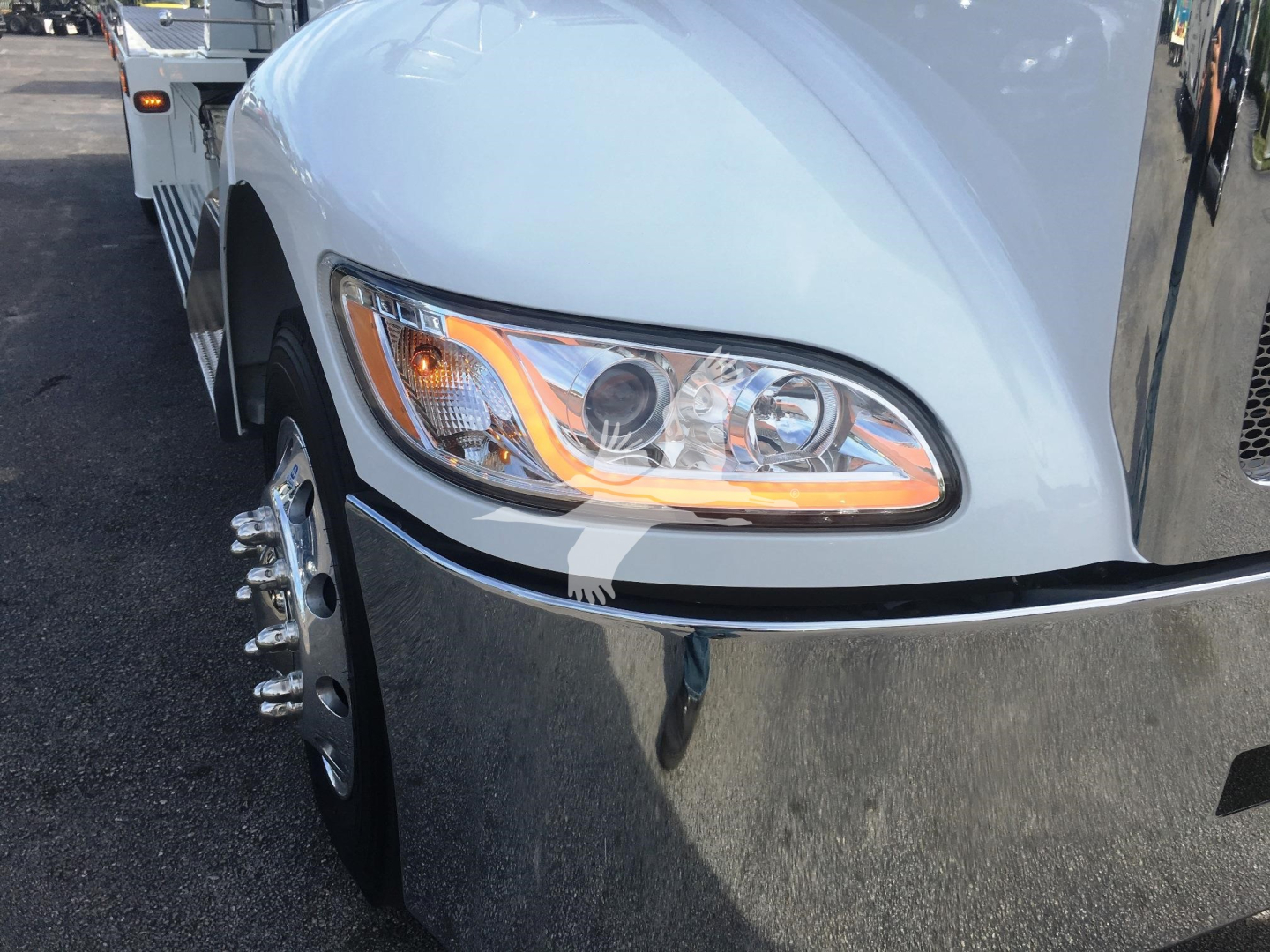Front left headlight area of a white 2022 Peterbilt 337 rollback truck with chrome bumper; wheel with chrome lug nuts visible.