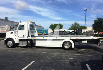 Flatbed tow truck in a parking lot, white cab and long white flatbed with towing equipment, Peterbilt 337 model, side view.