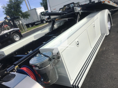 Peterbilt 337 rollback truck side view showing aluminum toolbox with lid, chrome air filter housing, and round canister with blue air hoses; rear wheel and part of flatbed visible.