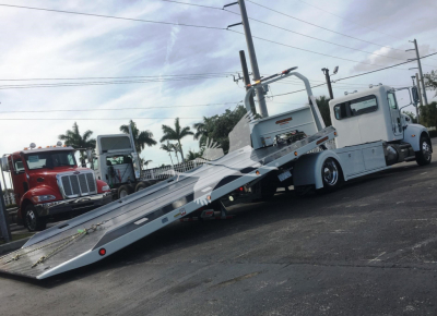 Peterbilt 337 rollback tow truck with a lowered steel ramp in the foreground and a white cab, parked lot with palm trees in the background.