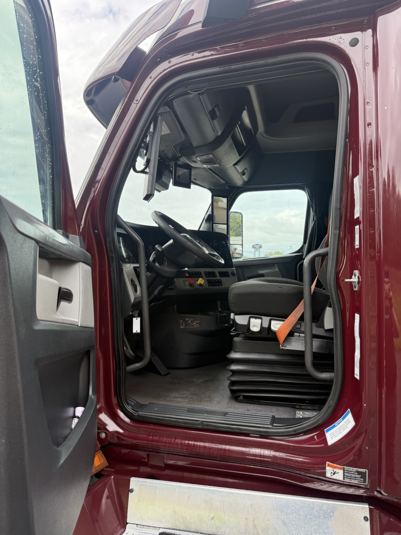 Truck cab interior view of a Freightliner Cascadia 126: driver seat, steering wheel, dashboard, and steps visible through open door.