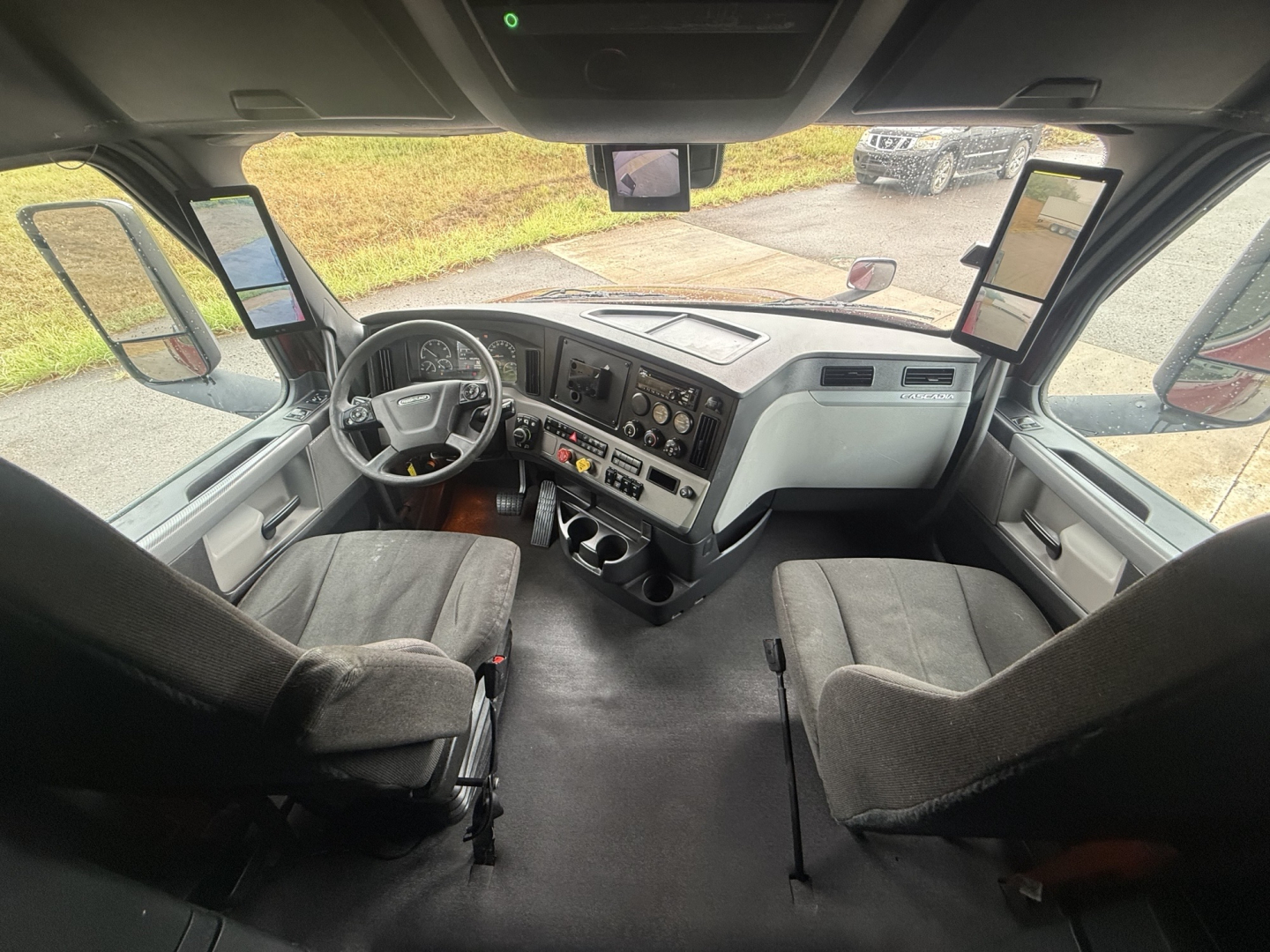 Cab interior of a Freightliner Cascadia truck showing steering wheel, dashboard with gauges, center console, two captain chairs, and side mirrors visible.
