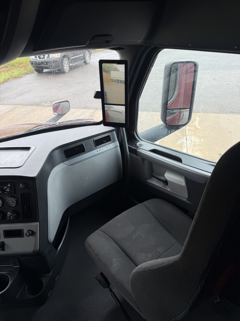 Truck cab interior view showing driver seat and dashboard with a dual side mirrors and a slim vertical mirror on the A-pillar. Text visible: none legible.