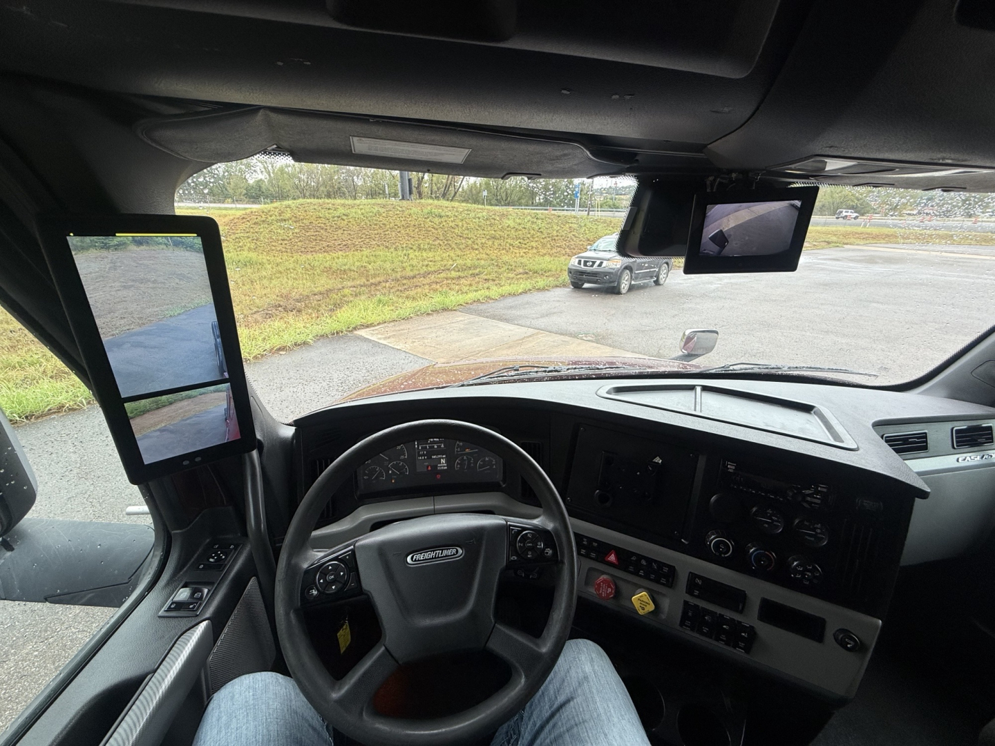 Cab interior of a Freightliner Cascadia 126 showing steering wheel, dashboard with gauges, two side mirrors, and a dash-mounted screen.