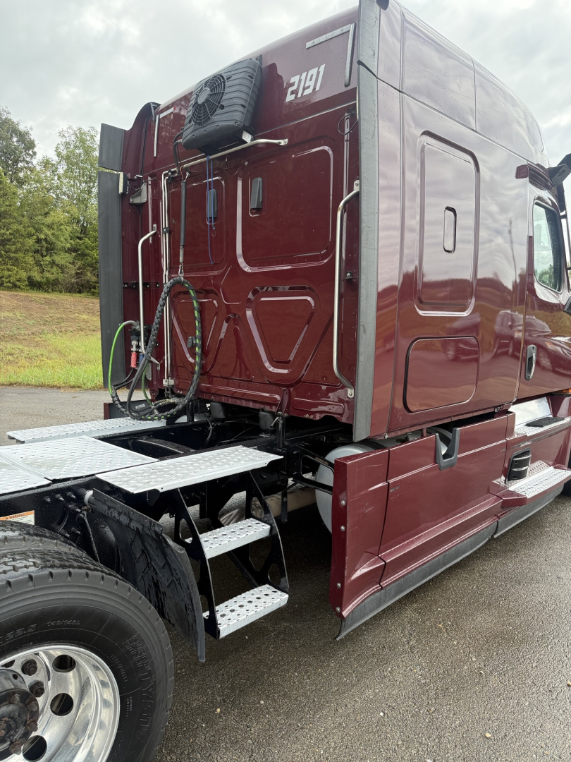 Rear quarter view of a maroon 2021 Freightliner Cascadia 126 truck showing side steps, rear cab area, spare tire, and hydraulic hoses. Visible features include a mounted exhaust and a ladder/steps.