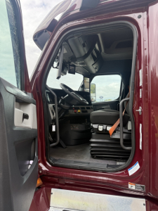 Truck cab interior view of a Freightliner Cascadia 126: driver seat, steering wheel, dashboard, and steps visible through open door.