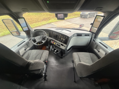 Cab interior of a Freightliner Cascadia truck showing steering wheel, dashboard with gauges, center console, two captain chairs, and side mirrors visible.