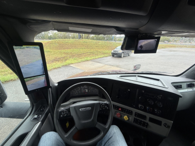 Cab interior of a Freightliner Cascadia 126 showing steering wheel, dashboard with gauges, two side mirrors, and a dash-mounted screen.