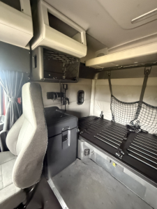 Cab interior of a Freightliner Cascadia126 sleeper showing bunk bed, storage cabinet, and seating area with overhead compartments.